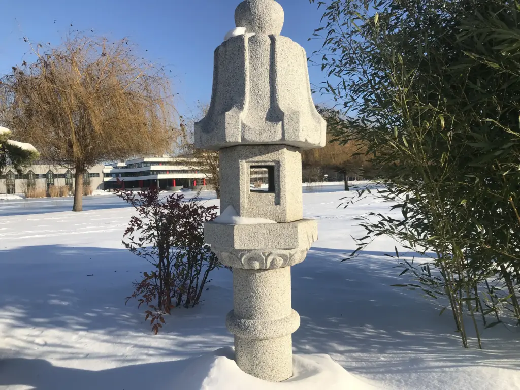 Hóval borított japánkertben álló kő lámpás, körülötte bambusz és téli növényzet.